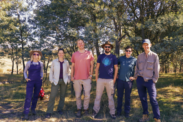 6 people standing side-by-side in nature, all smiling with their hands in their pockets.