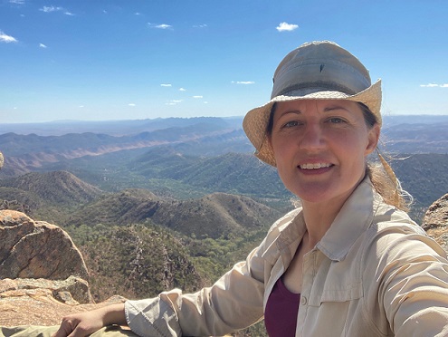Dr. Julie Groce wearing a beige hat and shirt smiling in the foreground atop a granite ridge with bushland in the background