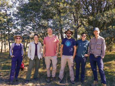 6 people standing side-by-side in nature, all smiling with their hands in their pockets.